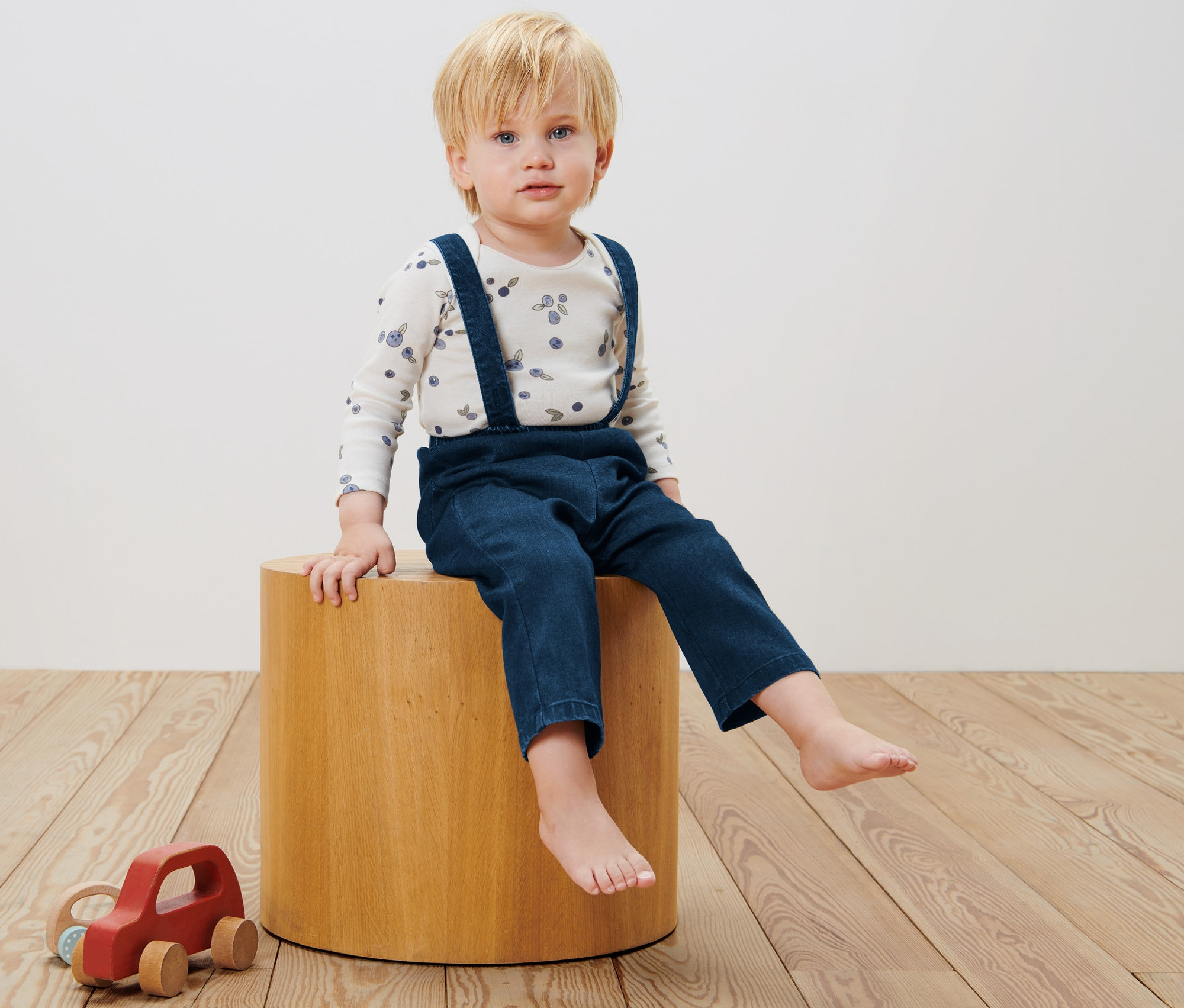Un garçon est assis sur un tabouret en bois et porte un pantalon en twill pour bébé avec une bavette.