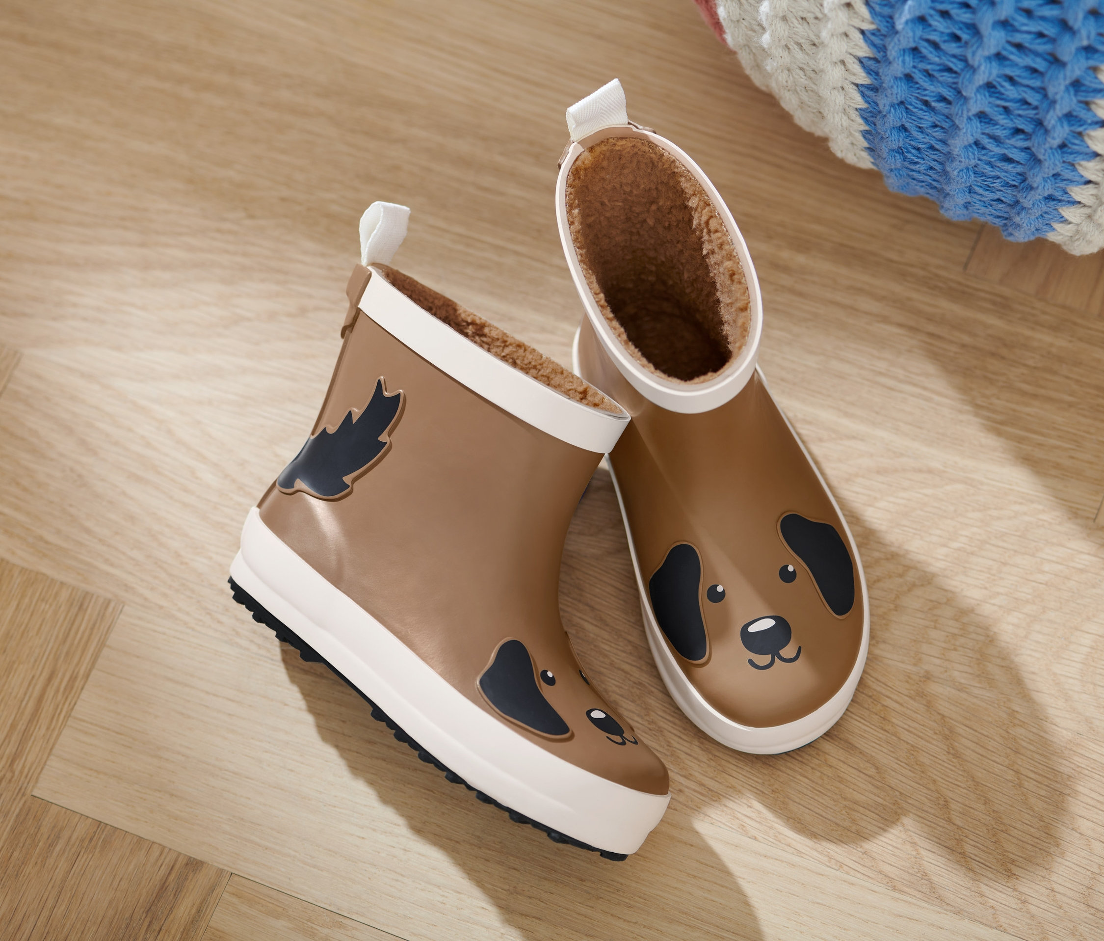 Deux bottes de pluie Chelsea pour enfant sur un plancher de bois. Une botte a une feuille et l'autre un chien.