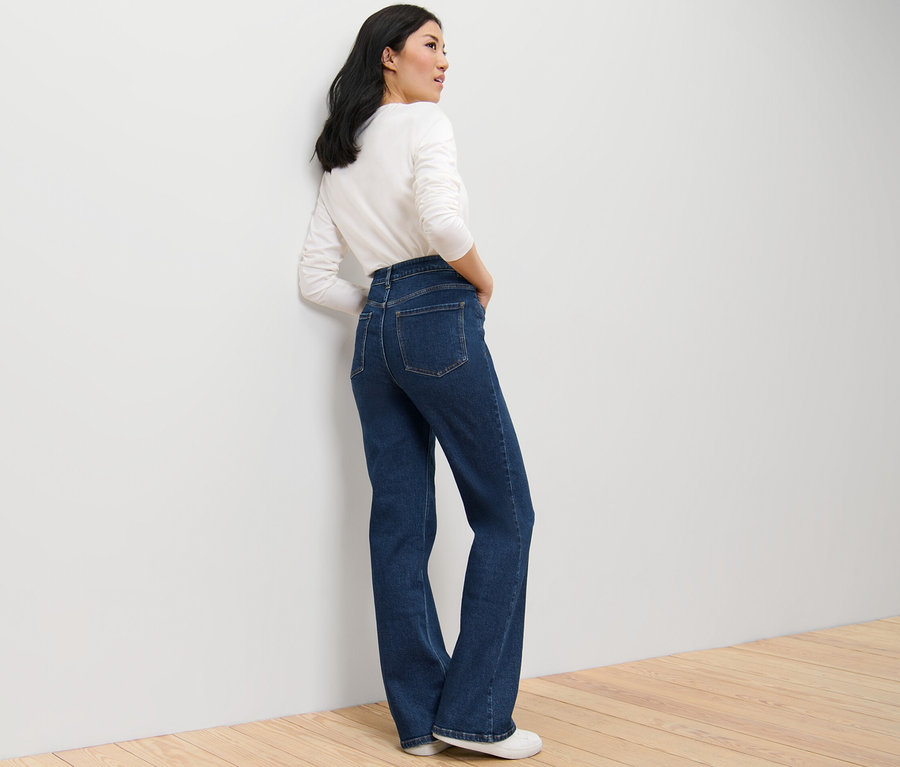 Une femme pose en jean bootcut – Fit « Lou » bleu foncé et un haut blanc à manches longues.