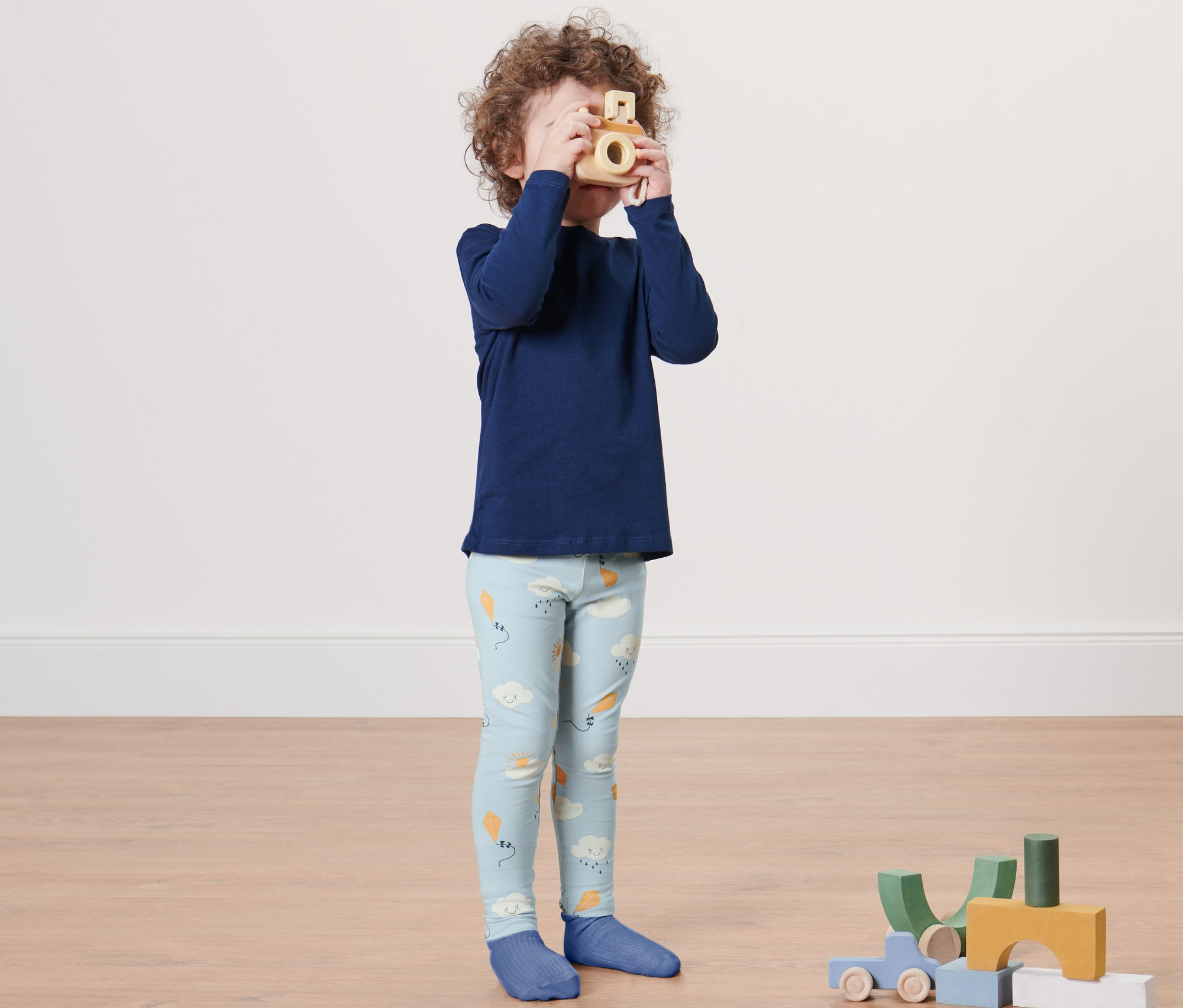 Un garçon avec un haut bleu et 2 leggings pour bébés et tout-petits joue avec un appareil photo en bois.