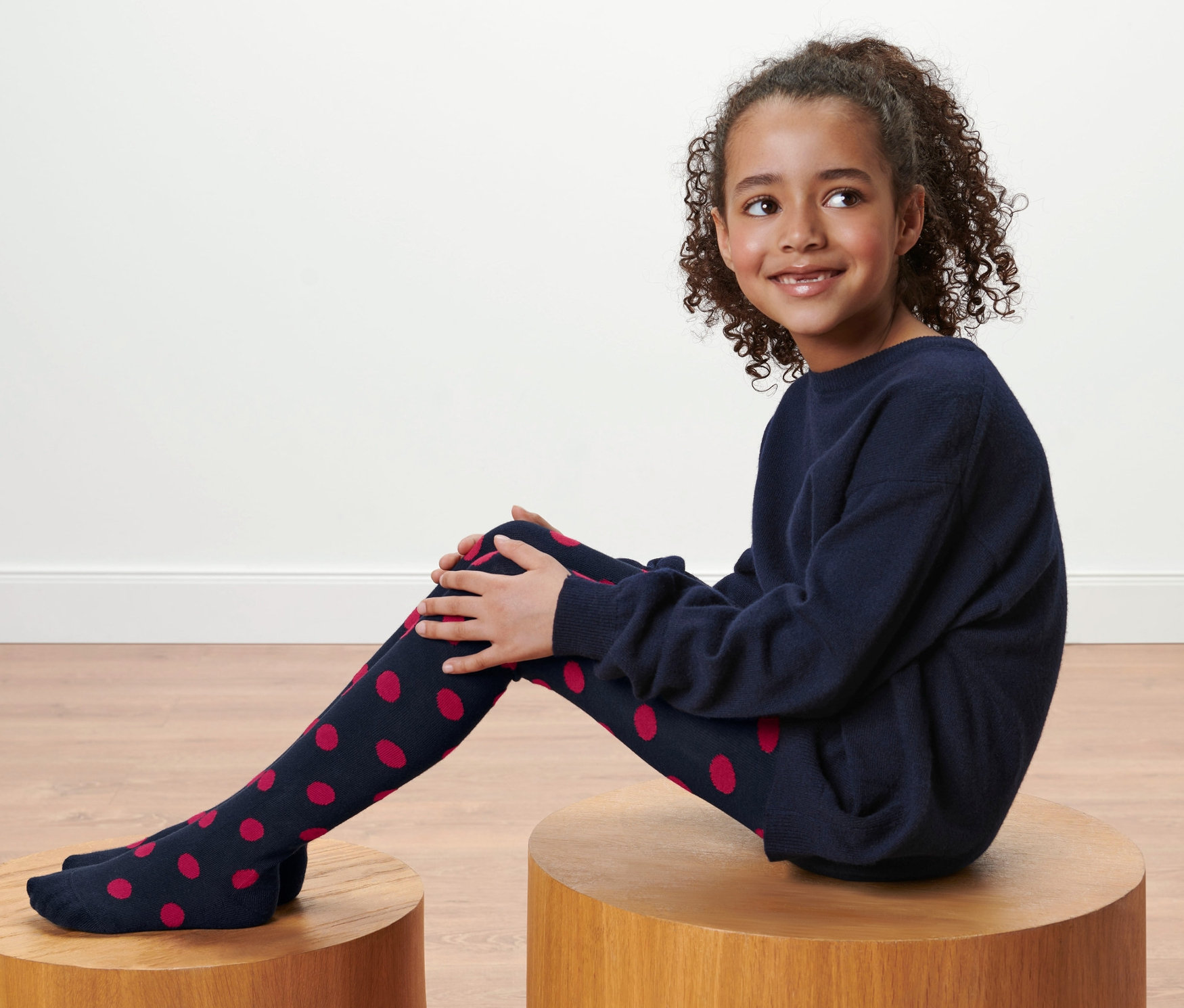 Une fille est assise sur un tabouret en bois et porte un collant bleu marine à pois rouges, 2	collants pour tout-petits.