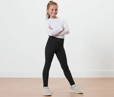 Une fille souriante pose, les bras croisés, portant un legging de sport pour enfants, un T-shirt blanc à manches longues et des baskets.