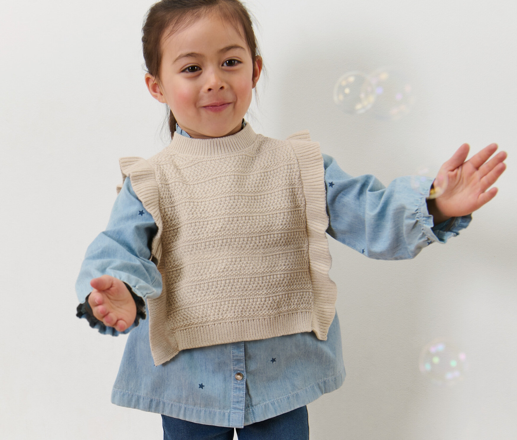 Fillette en pull sans manches en maille pour enfant posant, bras tendus.