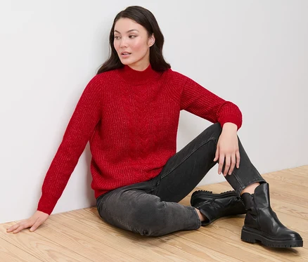 Une femme est assise sur un plancher en bois et porte un pull rouge en grosse maille à motif torsadé, un jean noir et des bottes noires.