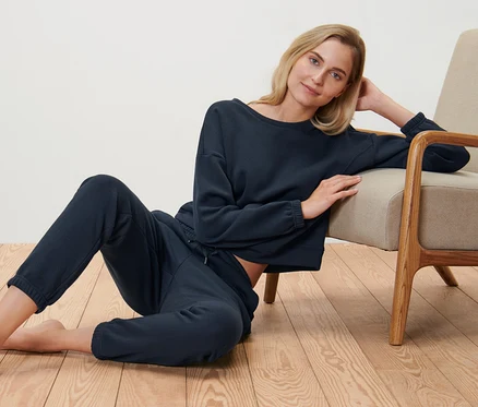Une femme blonde est assise sur le sol à côté d'un fauteuil et porte un survêtement d’intérieur bleu foncé.