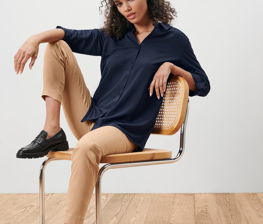 Femme assise sur une chaise portant un chemisier en jersey piqué bleu foncé et un pantalon beige.