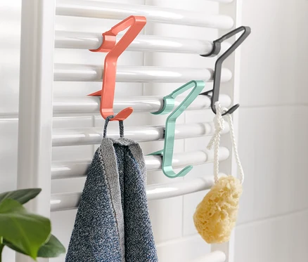 Sur un radiateur blanc sont accrochés 3 crochets à radiateur sur lesquels sont suspendus une serviette et une éponge.