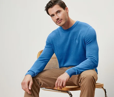 Un homme est assis sur une chaise et porte un pull bleu à encolure ronde.