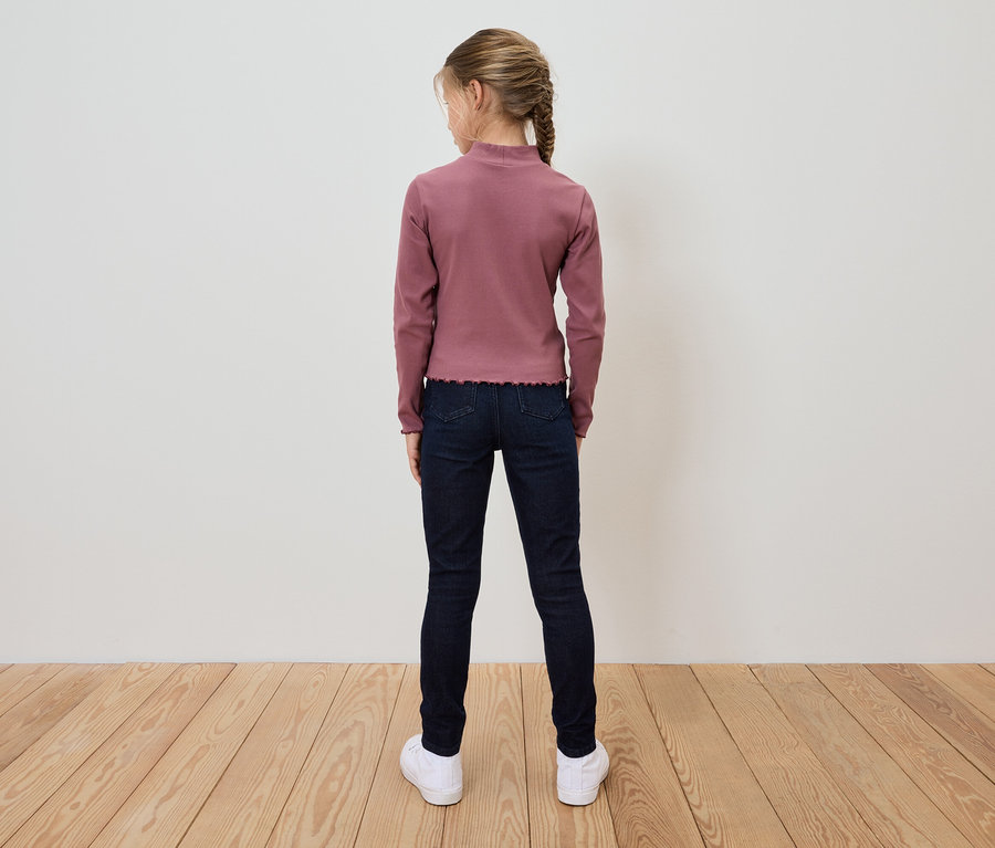 Une fille portant un T-shirt rose à manches longues, un jean bleu foncé et des chaussures blanches, se tient dos à la caméra.