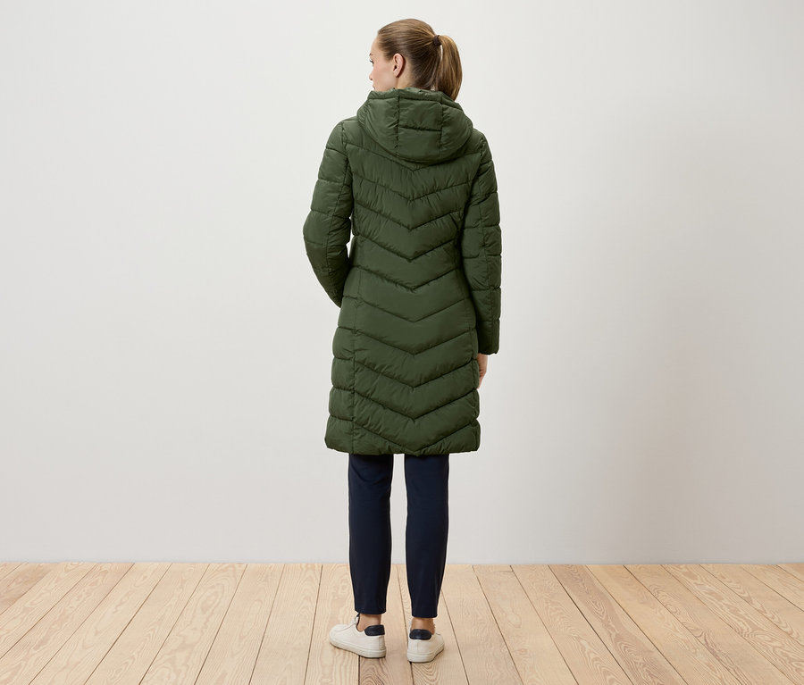 Une femme se tient dos à la caméra et porte un long manteau matelassé vert à capuche, un pantalon bleu marine et des baskets blanches.