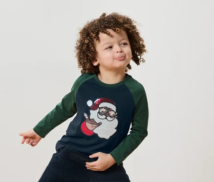 Un garçon aux cheveux bouclés porte un t-shirt bleu-vert avec une illustration du Père Noël.