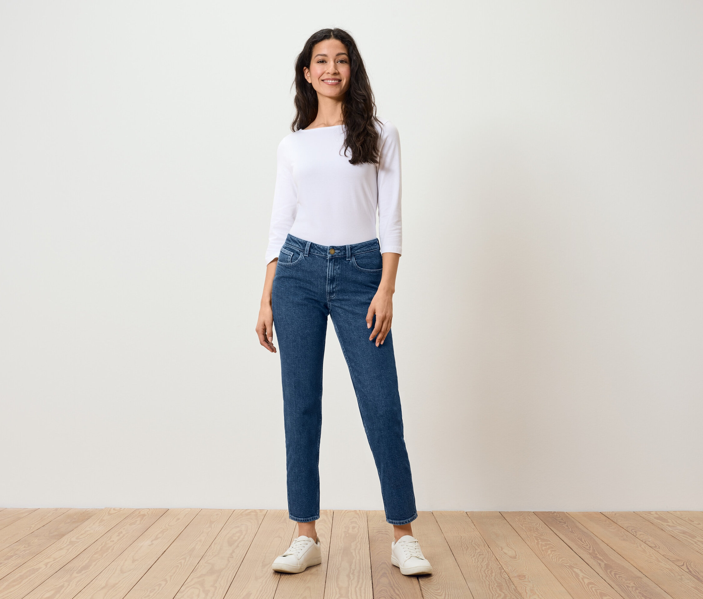 Une femme se tient sur un plancher en bois et porte un jean bleu, une chemise blanche et des baskets blanches.