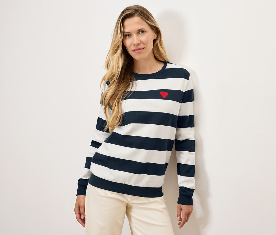 Blonde posant dans un pull rayé avec un cœur rouge et un pantalon clair.