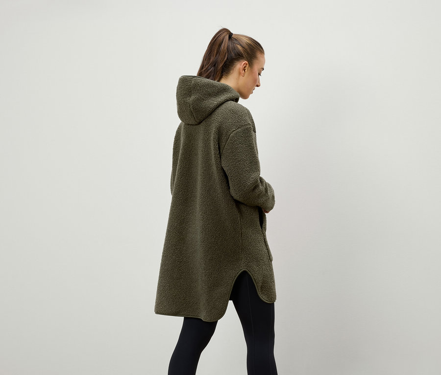 Une femme vue de dos porte un manteau à capuche en polaire vert olive et un legging noir.