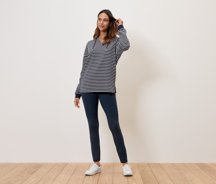 Une femme se tient sur un plancher en bois et porte un haut rayé, un legging bleu marine et des baskets blanches.