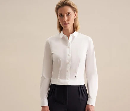Femme portant un chemisier Seidensticker Slimfit blanc avec un petit emblème foncé et un pantalon plissé foncé, sur fond clair.