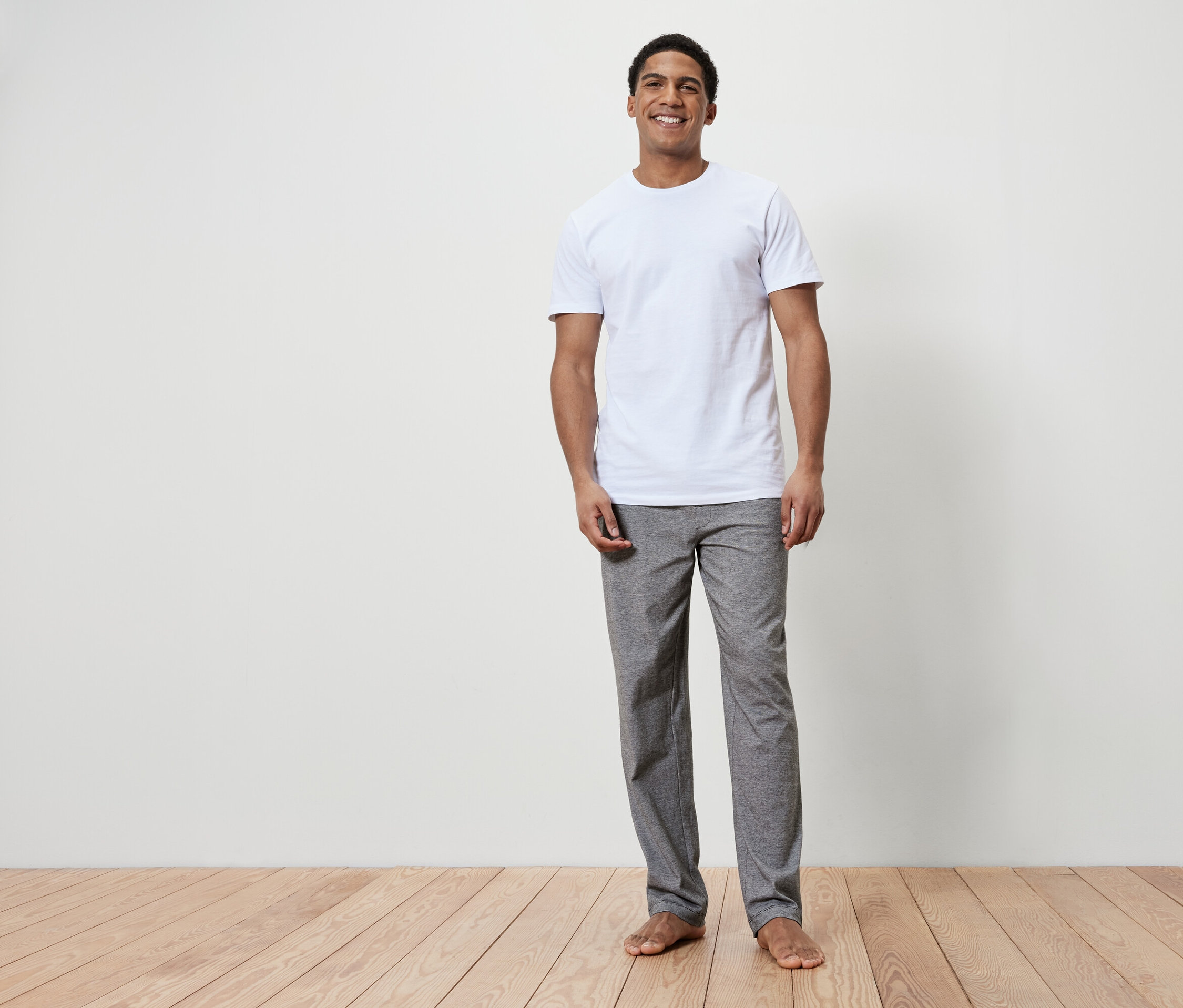 Un homme souriant se tient pieds nus sur un plancher en bois. Il porte un t-shirt blanc et un pantalon gris.