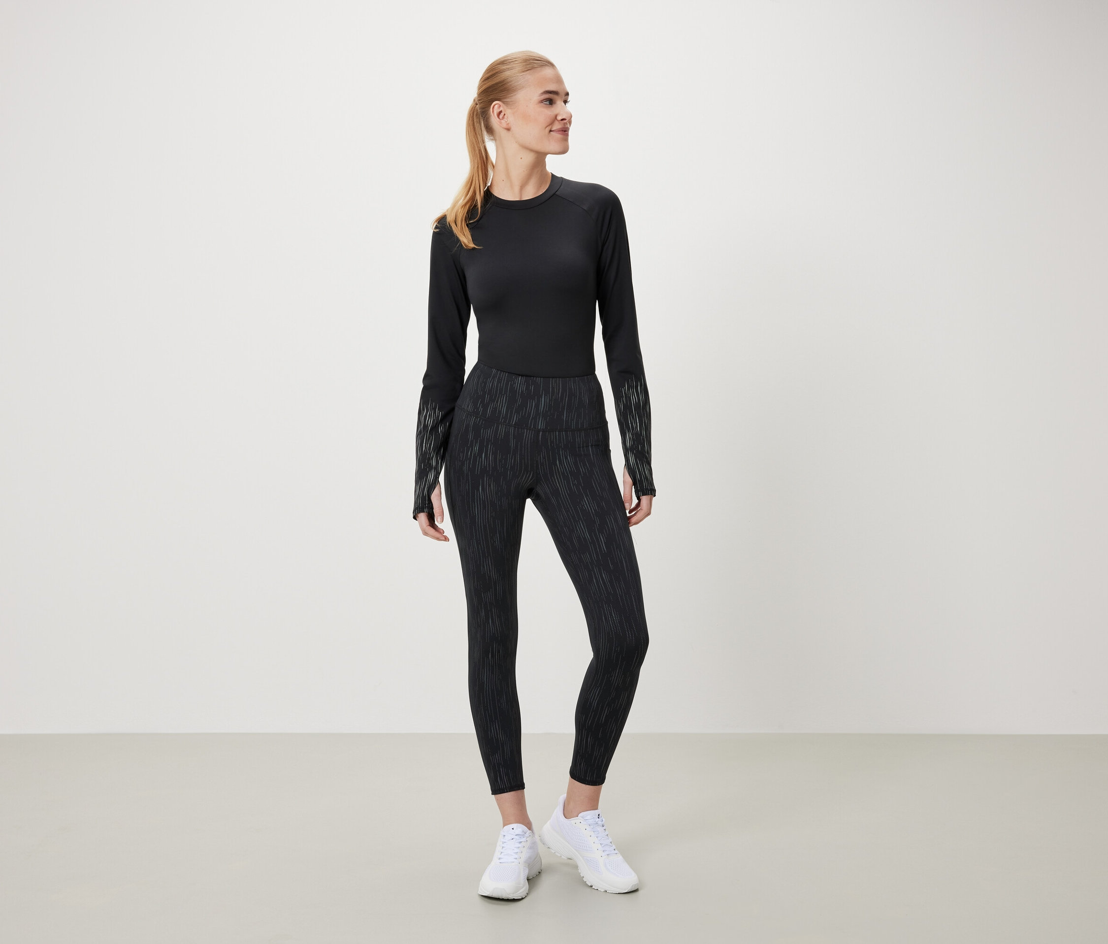 Une femme portant un haut noir à manches longues et un legging à motifs se tient sur un fond clair.