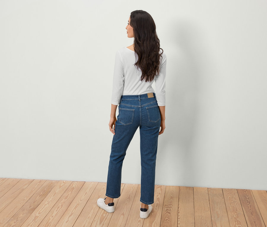 Femme aux longs cheveux bruns, dos à la caméra, portant un jean bleu et un haut blanc.