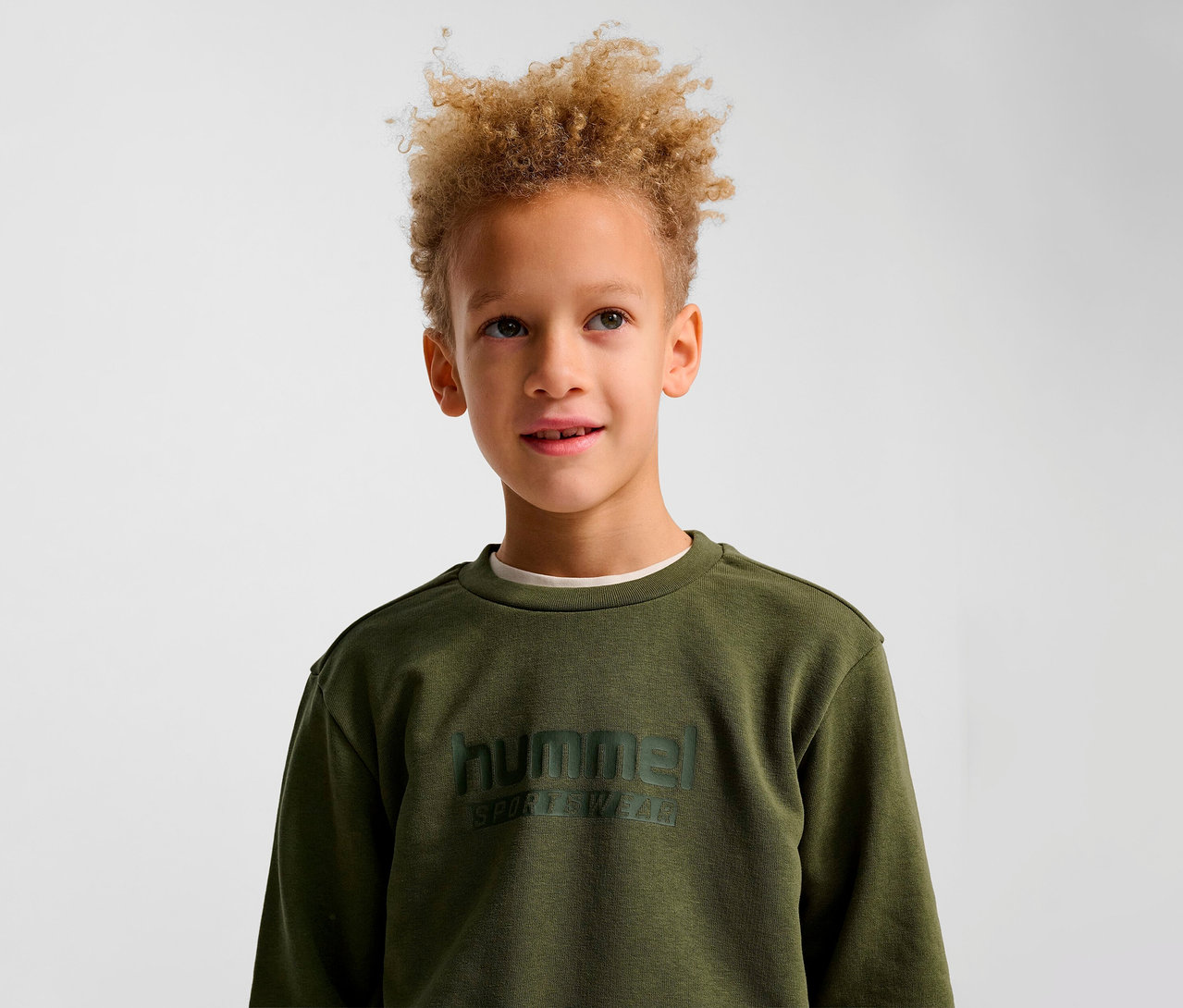 Un garçon aux cheveux blonds bouclés porte un sweat-shirt HUMMEL HMLJR Base vert.