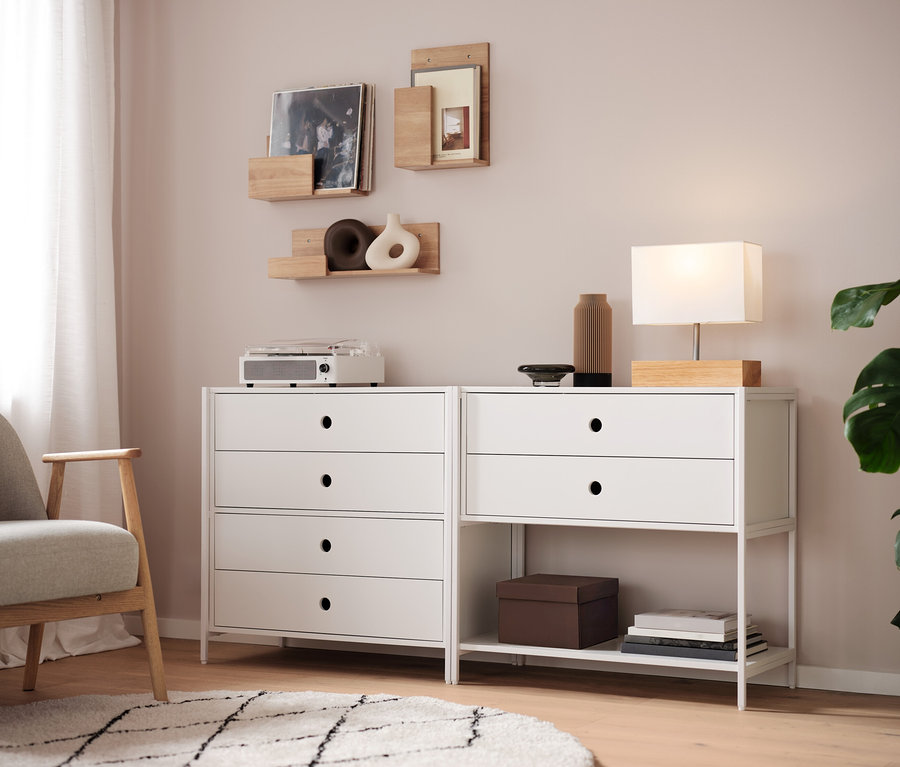 Deux commodes blanches sont placées l'une à côté de l'autre, avec une lampe, un vase et une radio dessus. Des étagères sont accrochées au mur.