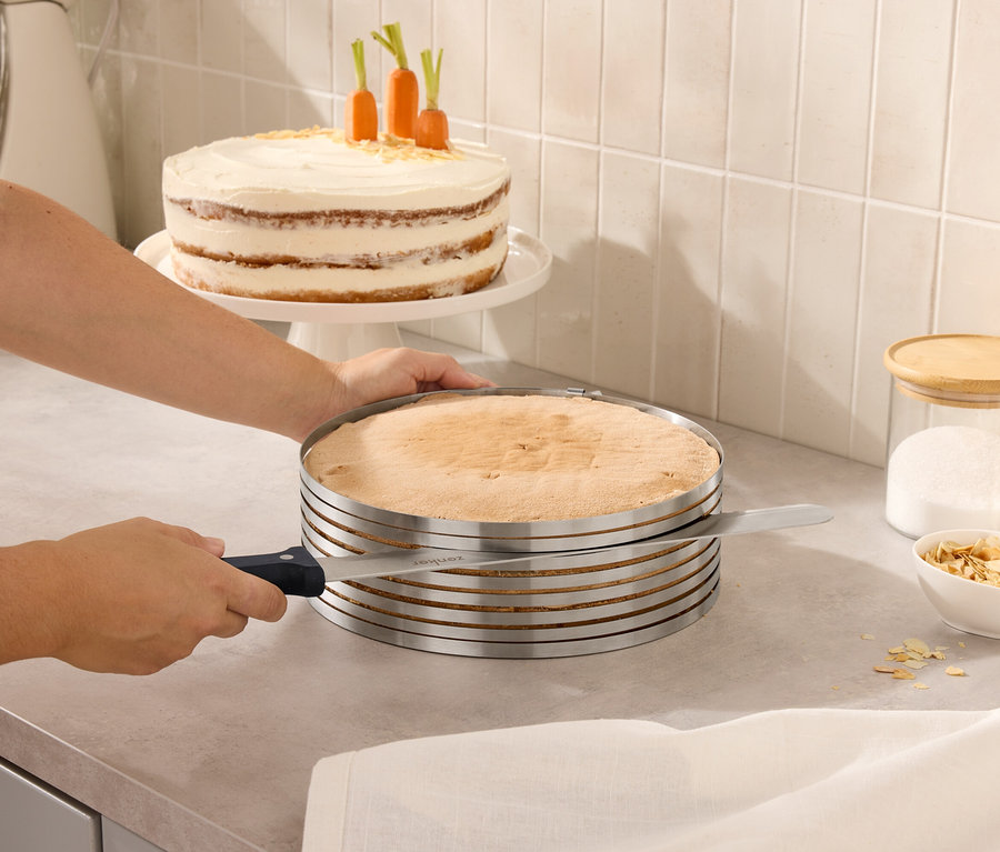 Une personne coupe un gâteau avec un coupe-gâteau réglable. En arrière-plan, il y a un gâteau avec des carottes et des bocaux avec des couvercles.