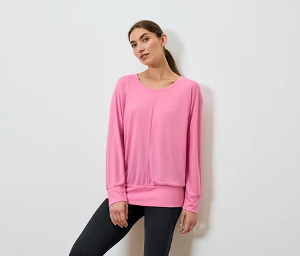 Une femme pose, portant un haut de sport rose à manches longues et un legging noir.