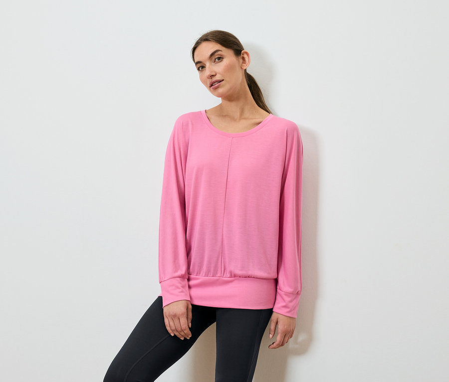 Une femme pose, portant un haut de sport rose à manches longues et un legging noir.