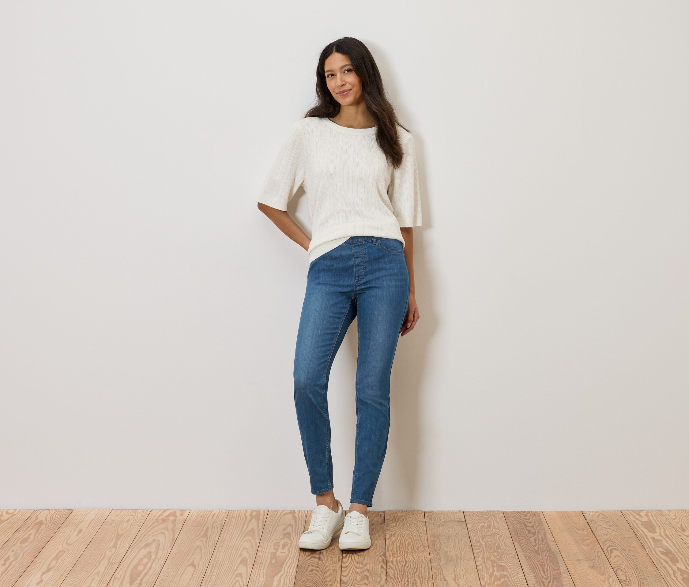 Une femme se tient devant un mur, portant un haut blanc, un jean bleu et des chaussures blanches.