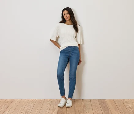 Une femme se tient devant un mur, portant un haut blanc, un jean bleu et des chaussures blanches.