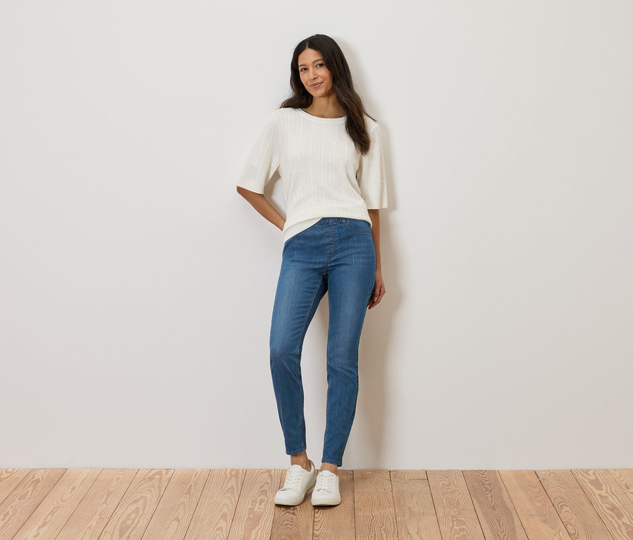 Une femme se tient devant un mur, portant un haut blanc, un jean bleu et des chaussures blanches.