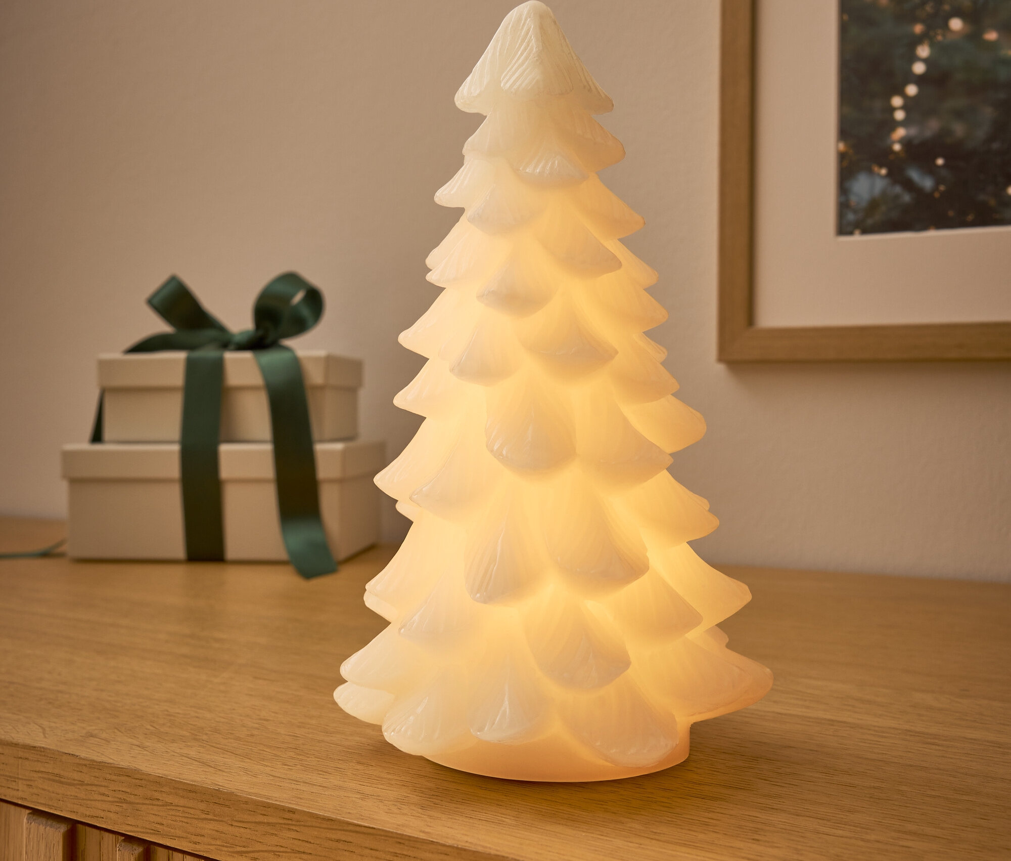 Sur une commode en bois, il y a un arbre lumineux, derrière lui deux cadeaux avec un ruban vert foncé et une image avec un motif de Noël.