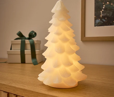 Sur une commode en bois, il y a un arbre lumineux, derrière lui deux cadeaux avec un ruban vert foncé et une image avec un motif de Noël.