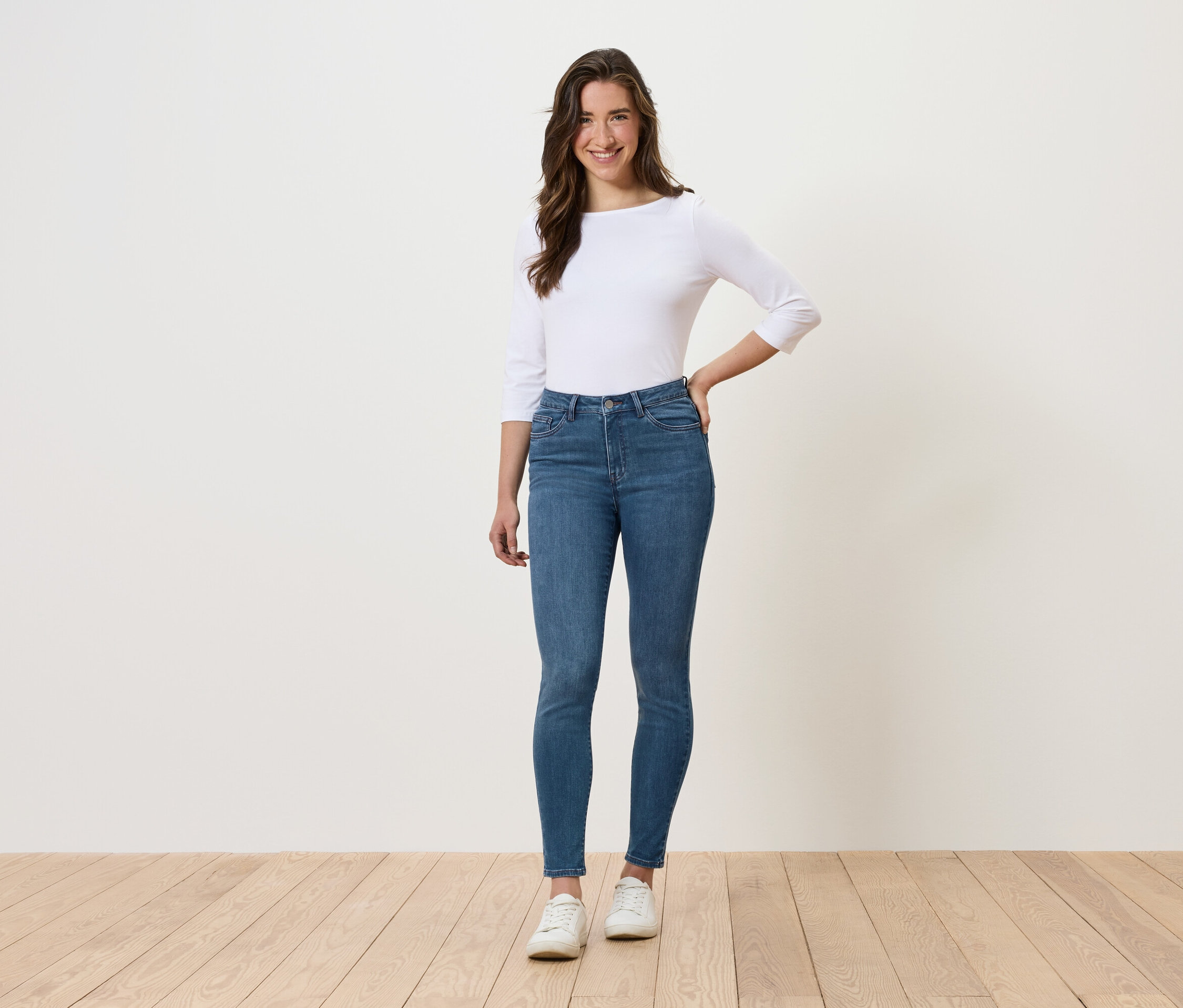 Une femme se tient debout et pose en jean bleu, haut blanc et chaussures blanches.