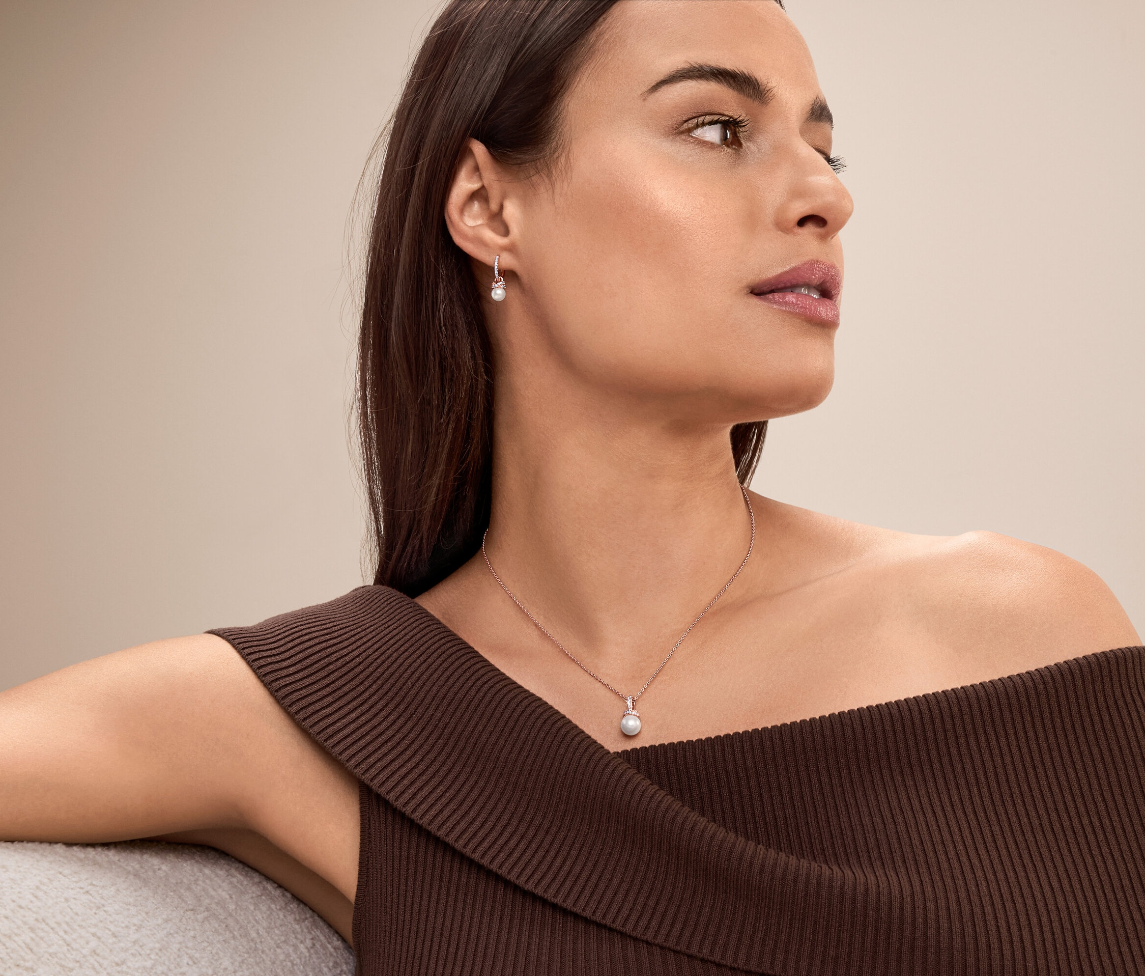 Une femme aux cheveux bruns et au haut marron à épaules dénudées portant un collier et des boucles d'oreilles en perles.