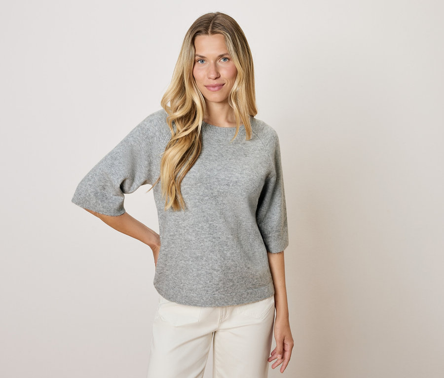 Une femme blonde portant un pull gris à manches trois-quarts et un pantalon blanc pose sur un fond clair.