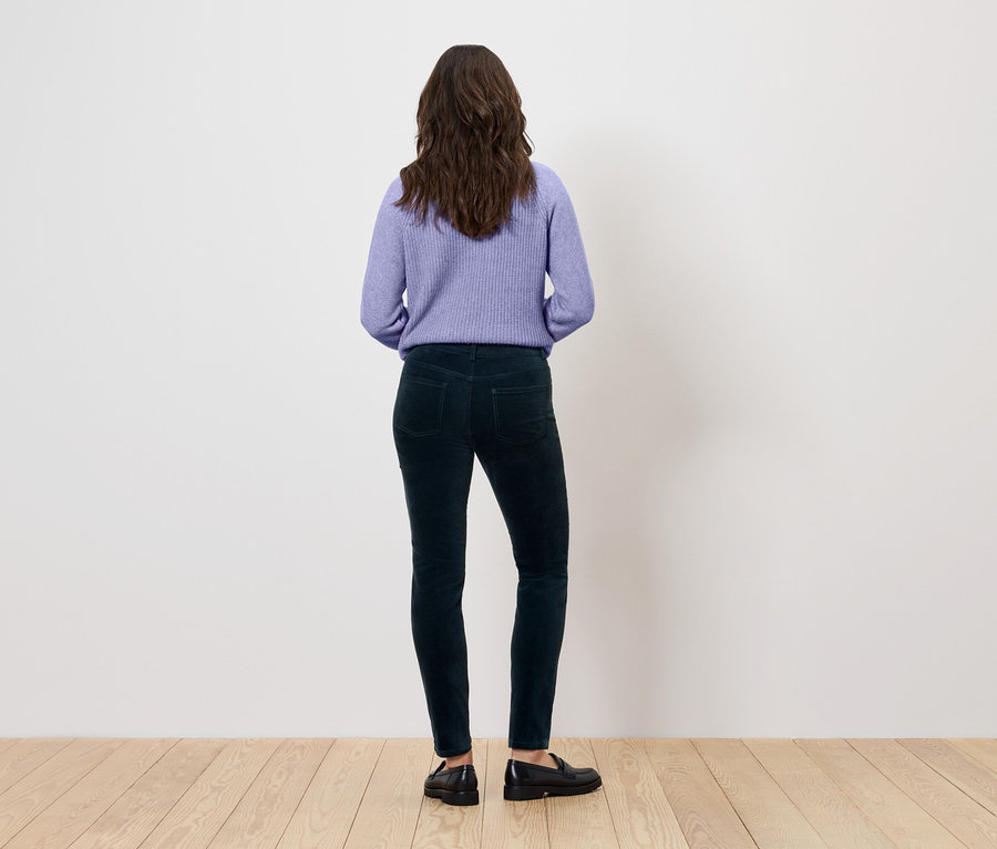 Vue arrière d'une femme aux cheveux bruns ondulés portant un pull violet, un pantalon vert foncé et des chaussures noires.