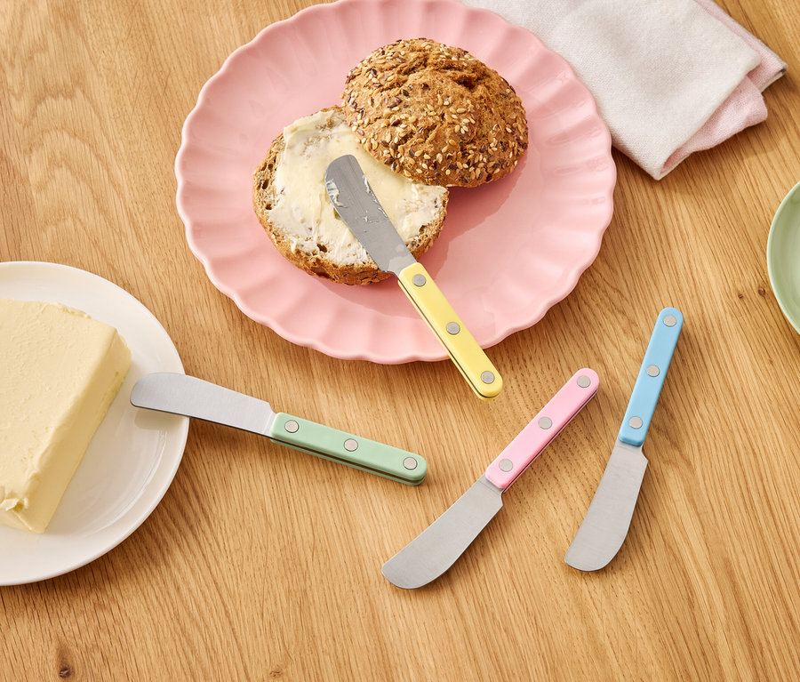 Un pain au beurre sur une assiette rose à côté d'une motte de beurre et de couteaux colorés sur une table en bois.