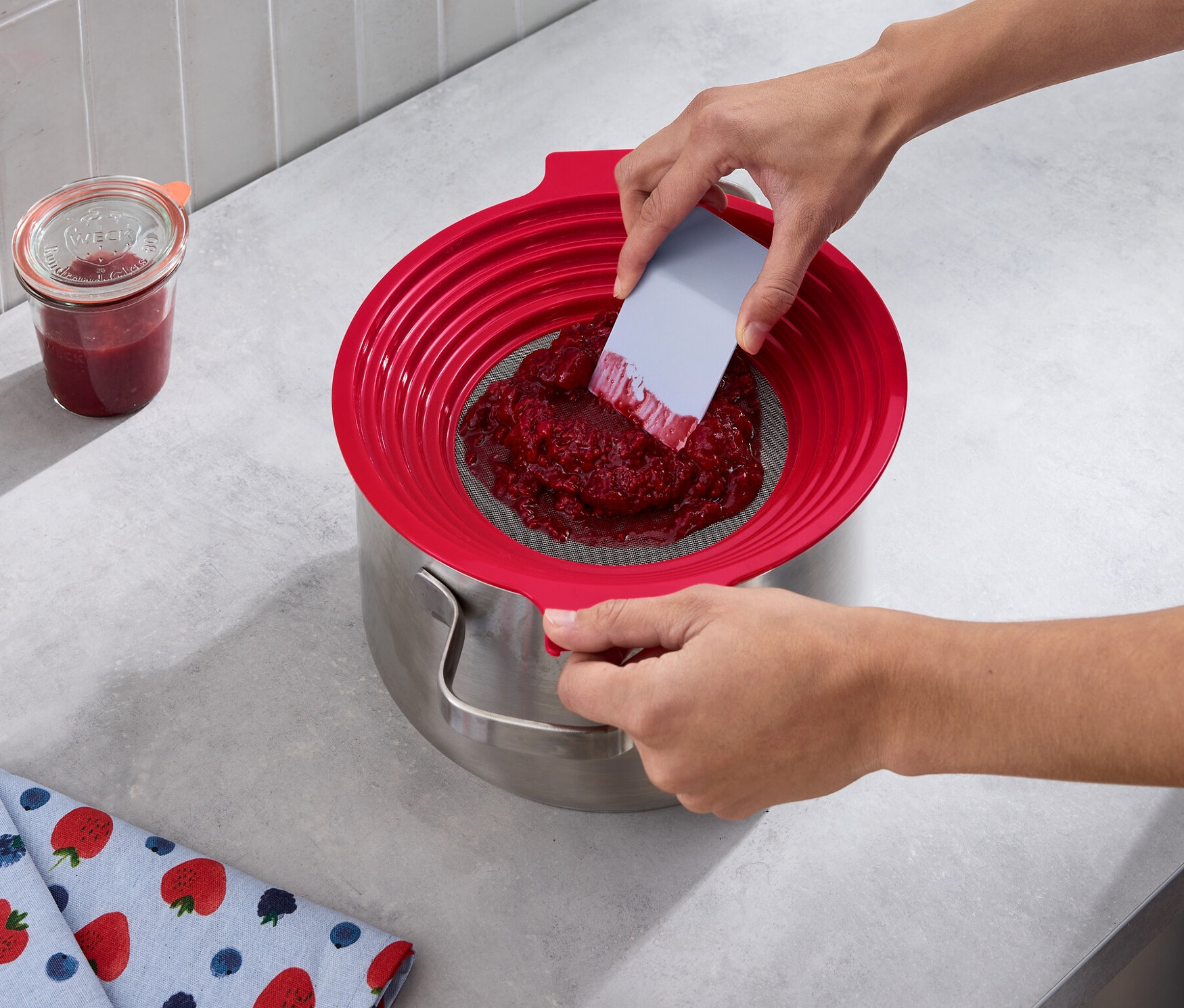 Une personne passe des fruits rouges dans une passoire pour farine et pour tamiser au-dessus d'une casserole. à cÎté se trouvent un pot de confiture et un torchon avec des motifs de fraises.