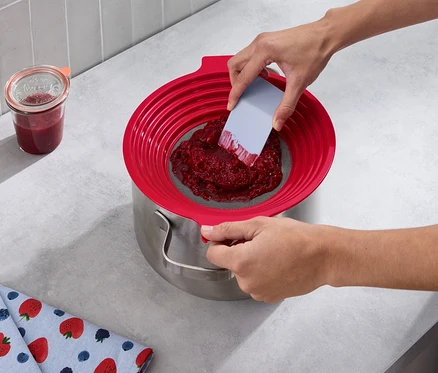 Une personne passe des fruits rouges dans une passoire pour farine et pour tamiser au-dessus d'une casserole. À côté se trouvent un pot de confiture et un torchon avec des motifs de fraises.