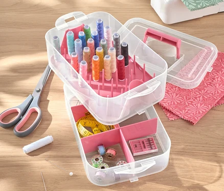 Boîte à outils ouverte en plastique avec fils, ciseaux, aiguilles et machine à coudre sur une table.