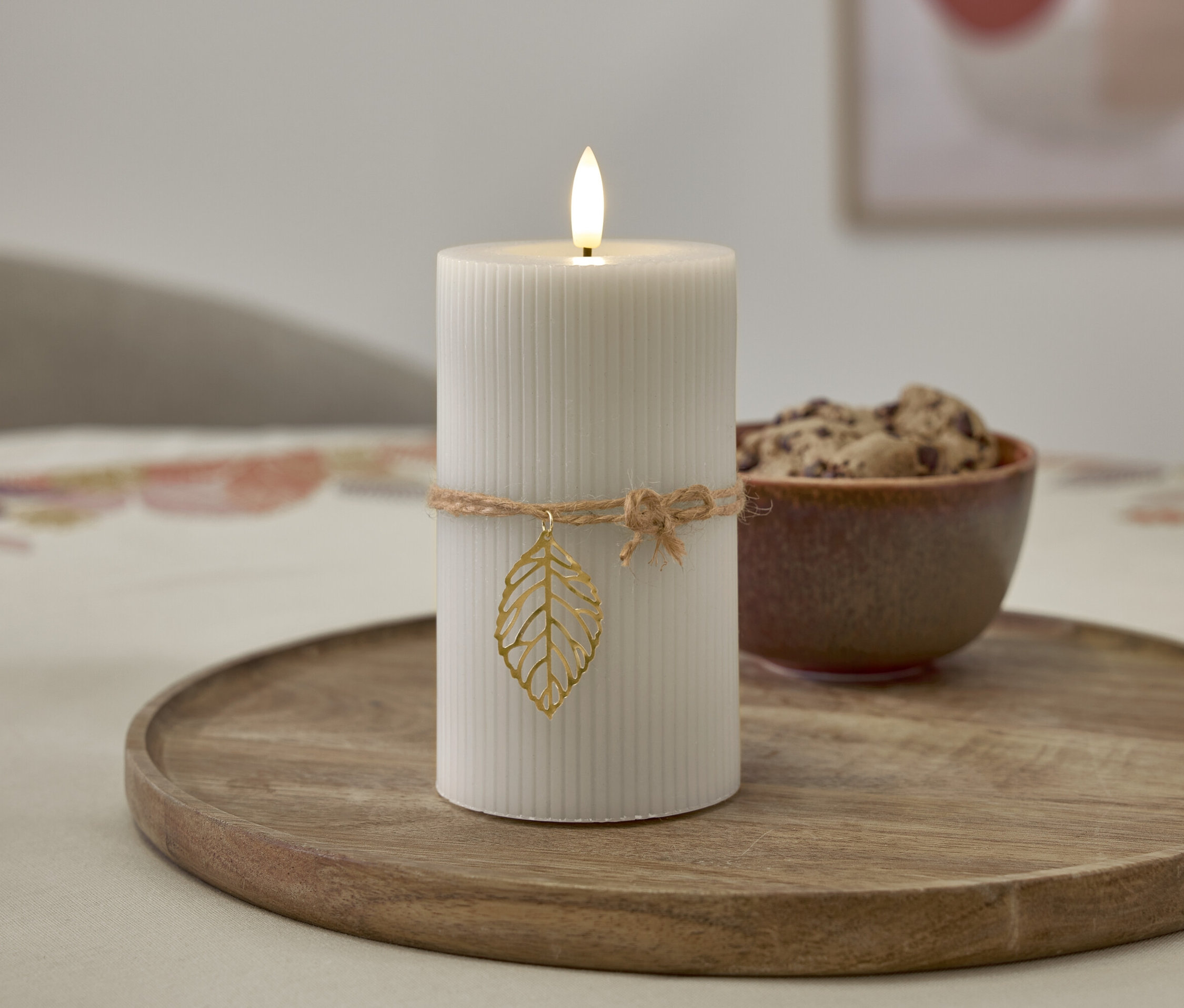 Sur un plateau en bois se trouve une bougie blanche avec une flamme, entourée d'une ficelle avec une décoration en forme de feuille, à côté d'un bol de biscuits.