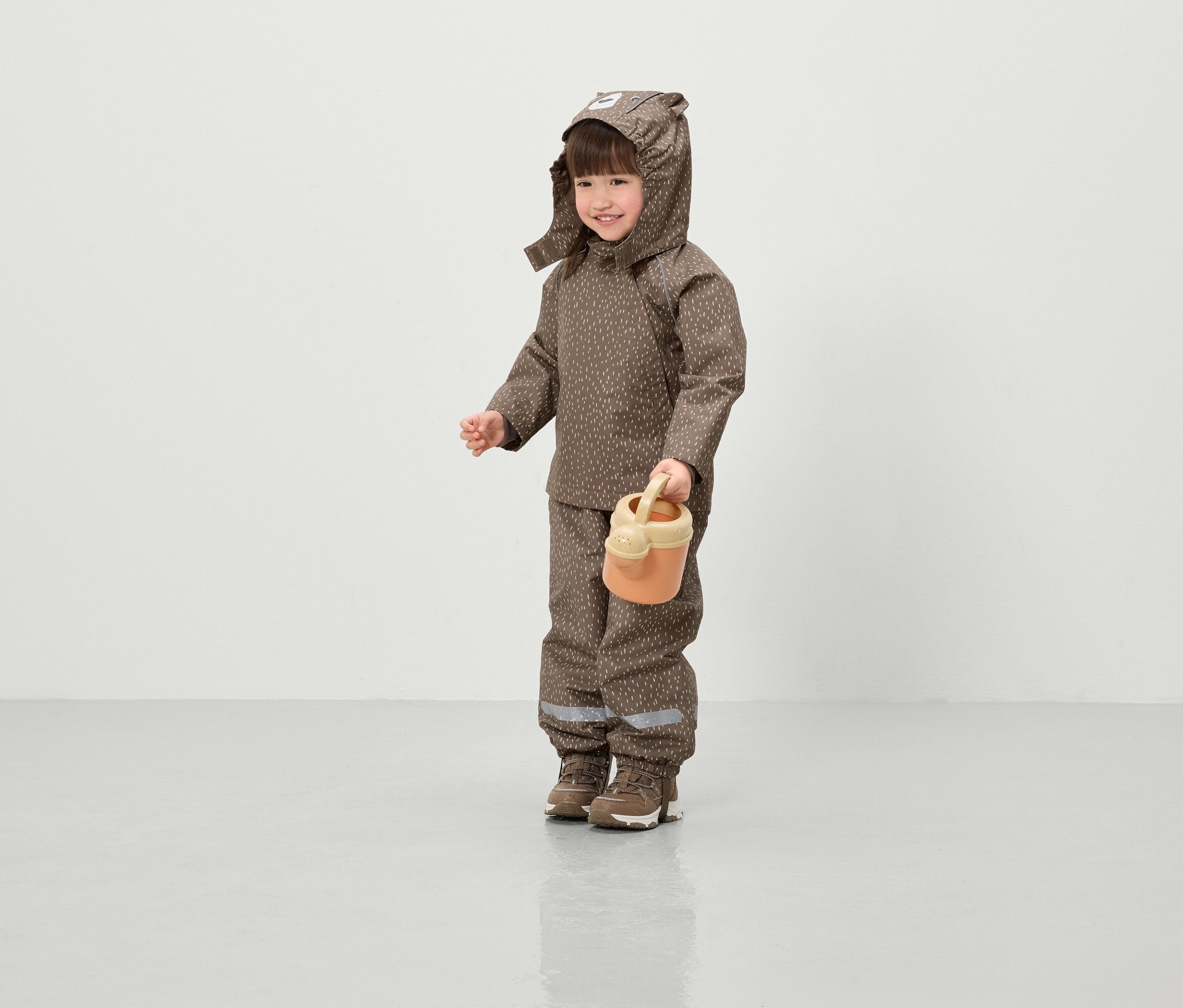 Un enfant souriant vêtu d'une veste et d'un pantalon marron à motifs avec capuche tient un arrosoir orange.