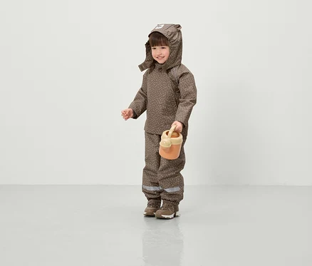 Un enfant souriant vêtu d'une veste et d'un pantalon marron à motifs avec capuche tient un arrosoir orange.