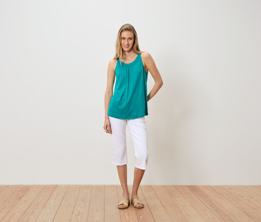 Une femme se tient devant un mur blanc, portant un débardeur turquoise, un pantalon capri blanc et des sandales.
