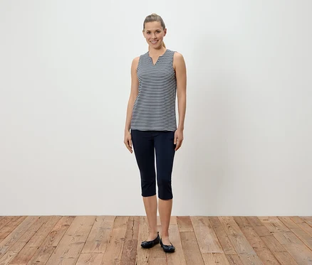 Une femme se tient devant un mur blanc et porte un débardeur rayé bleu et blanc, un pantalon capri bleu et des ballerines.