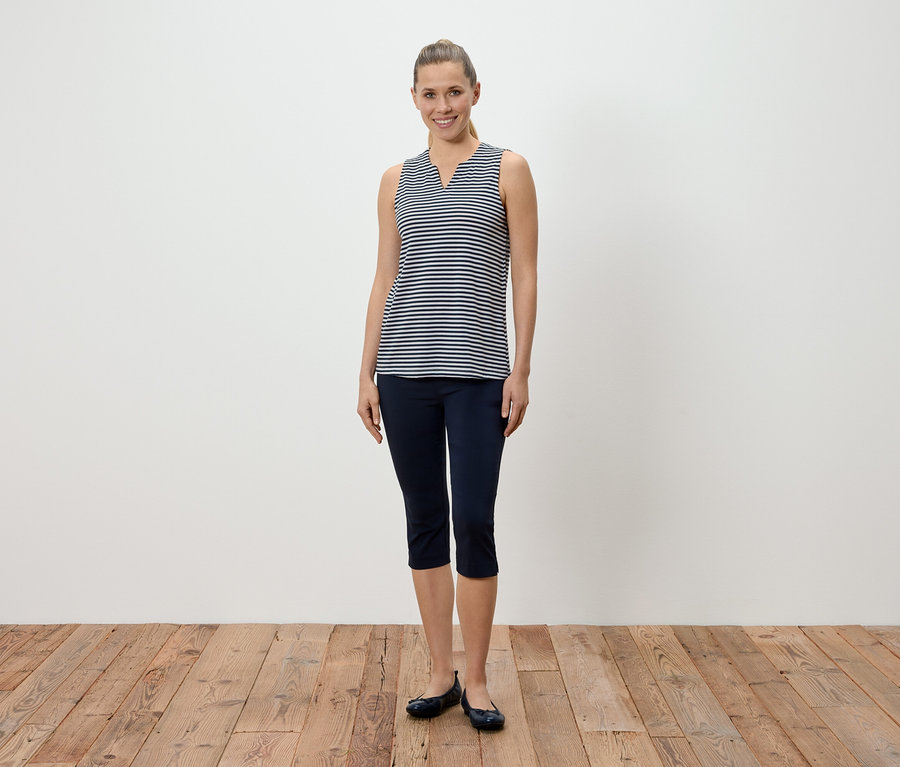Une femme se tient devant un mur blanc et porte un débardeur rayé bleu et blanc, un pantalon capri bleu et des ballerines.