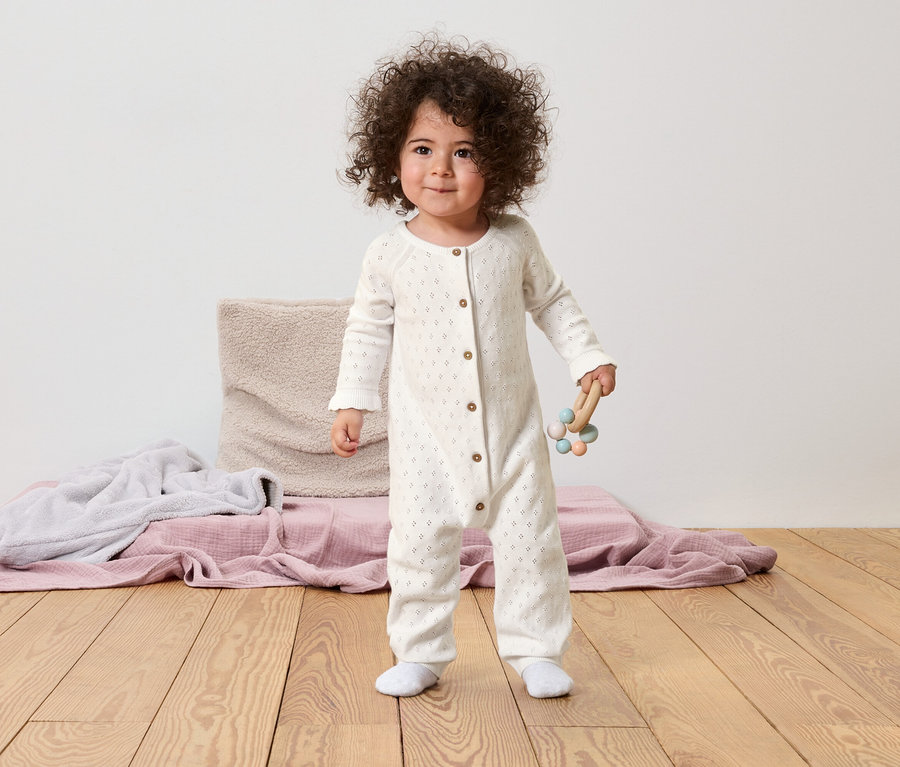 Un jeune enfant aux cheveux bouclés se tient sur un plancher en bois et tient un jouet en bois dans sa main. L'enfant porte un pyjama blanc.