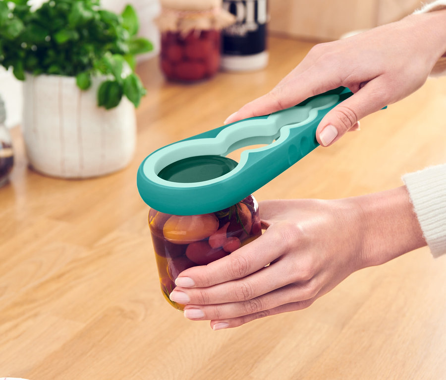 Une personne ouvre un bocal avec un ouvre-bocal turquoise et vert sur un comptoir de cuisine en bois.