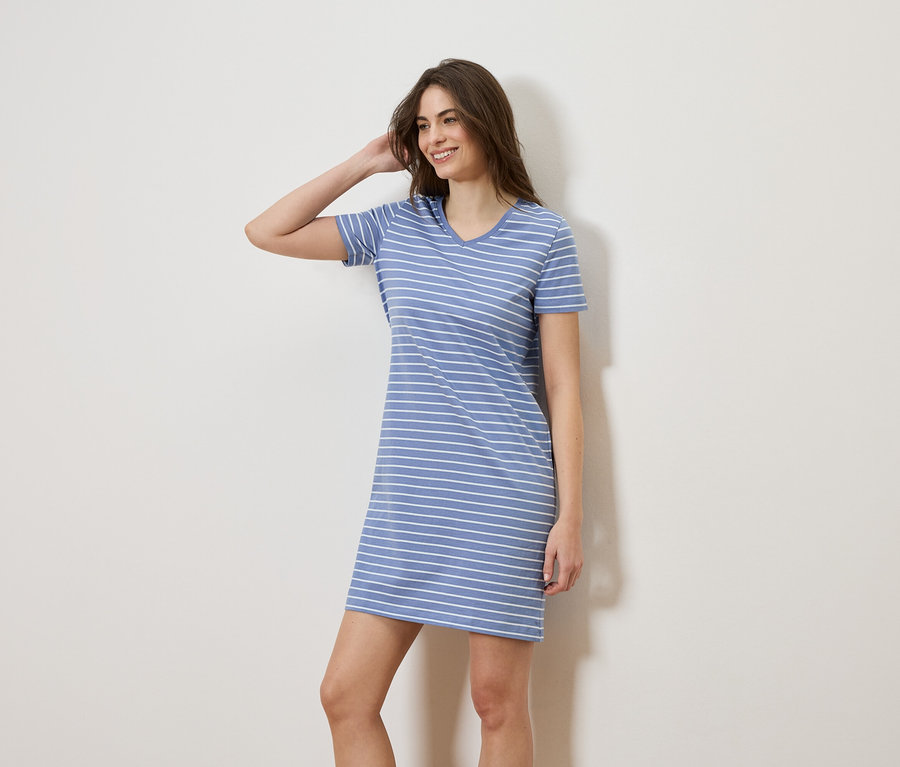 Une femme aux cheveux bruns pose dans une robe t-shirt rayée bleu et blanc devant un mur blanc.